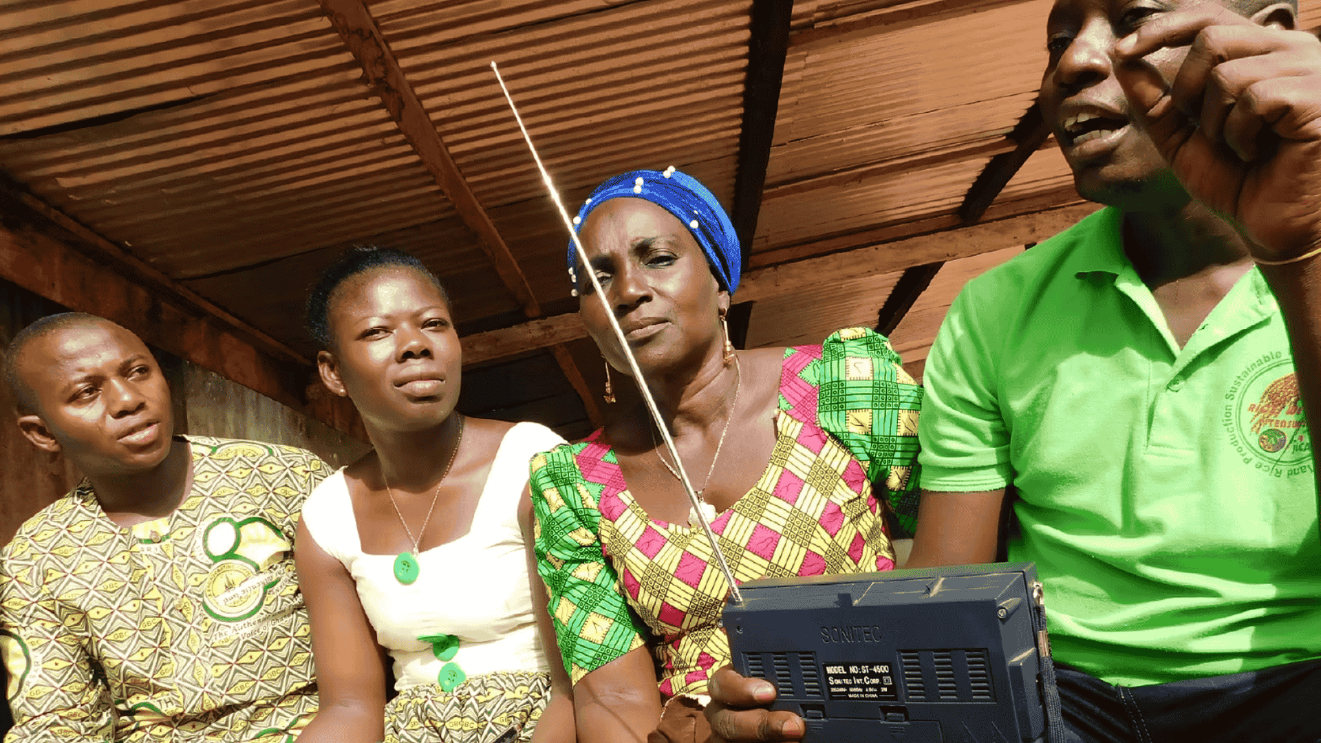 Group of farmers in colorful clothing sitting together and listening to a portable radio, highlighting community storytelling and farmer engagement.