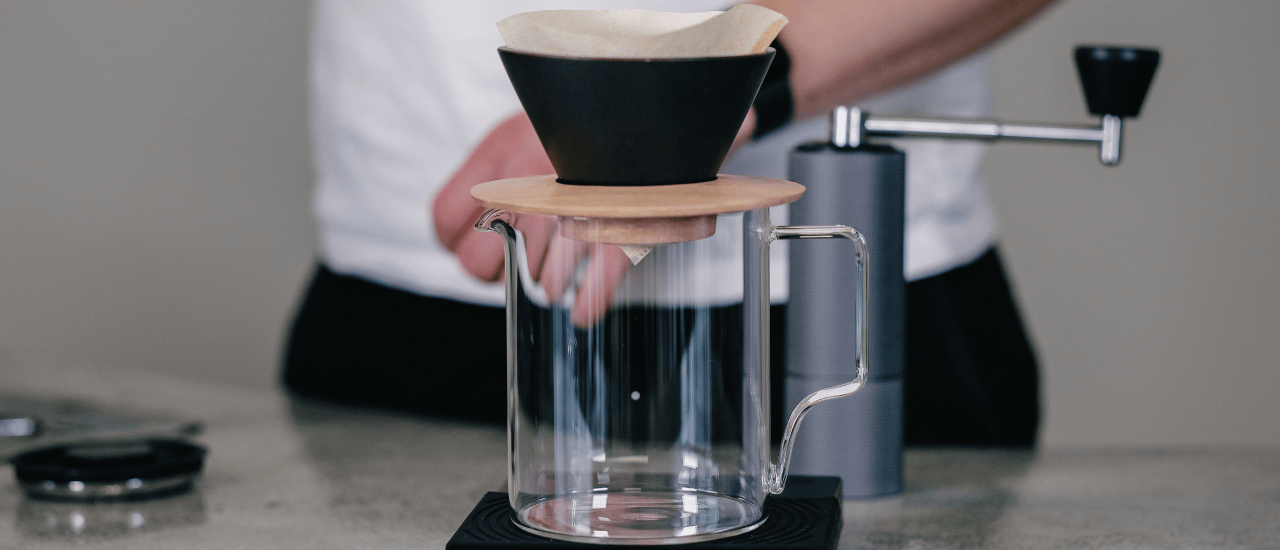 A glass carafe with a black V60 dripper and paper filter set up for brewing coffee, with a manual coffee grinder in the background.