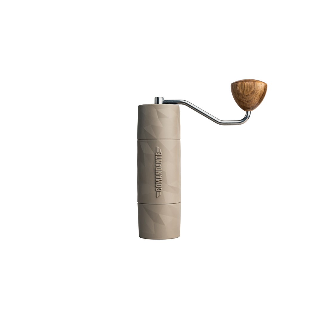 A beige Comandante hand coffee grinder with a textured, geometric finish. It features a metal crank arm with a wooden handle, extending from the top side of the grinder. The Comandante logo is embossed vertically on the body. The background is plain white.