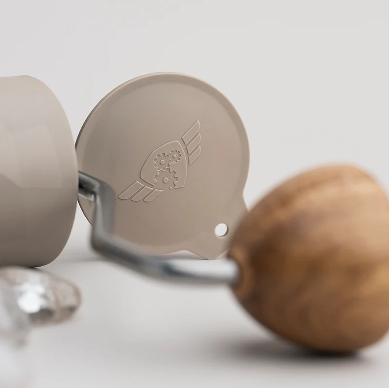 A close-up of the crank and internal components of a beige Comandante hand coffee grinder. The wooden handle is prominently in the foreground, with the metal crank arm leading to the grinder’s lid, which features an embossed gear-and-wings logo. The surface finish is matte, and the background is softly lit in neutral tones.