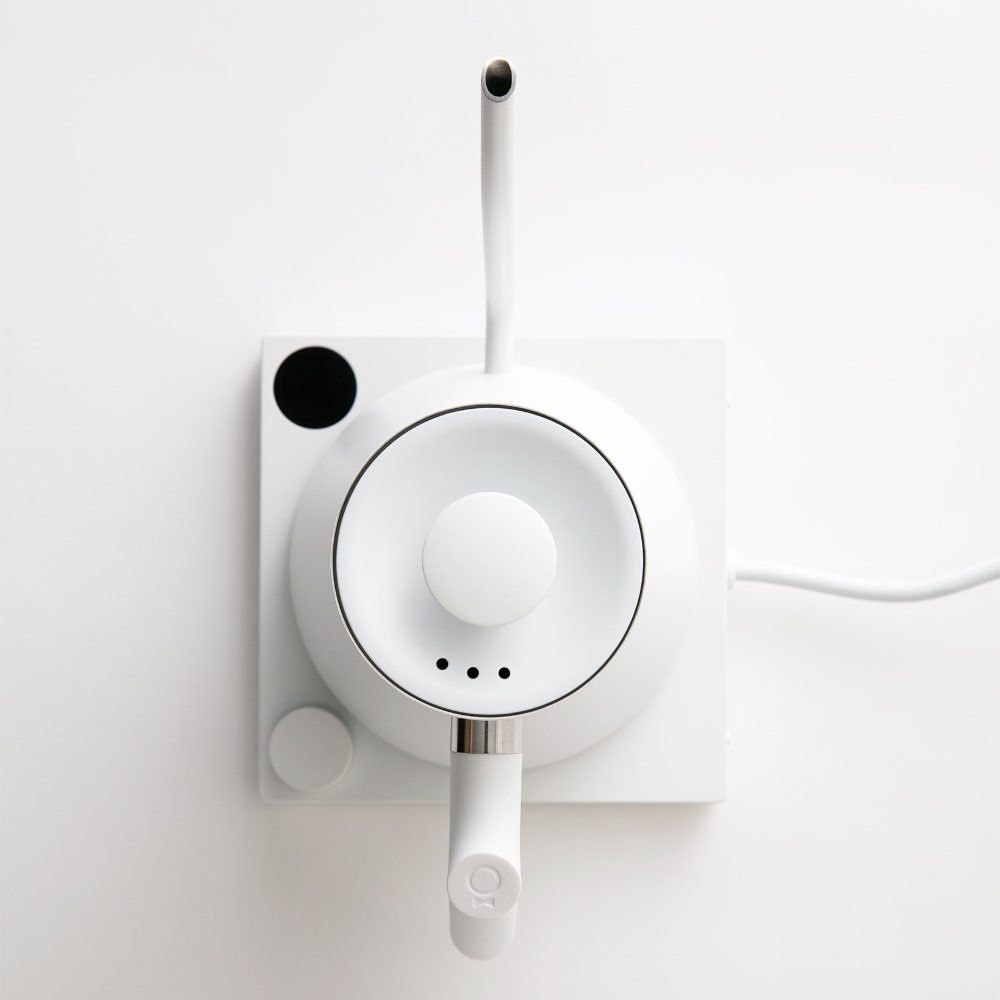Top-down view of a white Fellow Stagg EKG electric kettle on its square base. The kettle features a minimalist design with a gooseneck spout, a central round lid, and a cylindrical handle. The base includes a circular control knob and simple dot indicators. The power cord is visible extending from the right side. The setup is photographed against a clean white background.