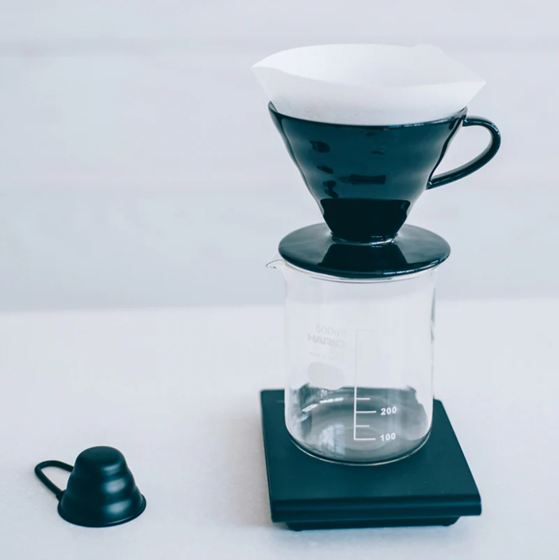 A black ceramic V60 coffee dripper with a white paper filter sits atop a glass Hario beaker marked with volume measurements. The setup is placed on a digital scale with a matte black finish. To the left, a black V60 measuring scoop rests on the countertop. The scene is set against a light, minimal background.