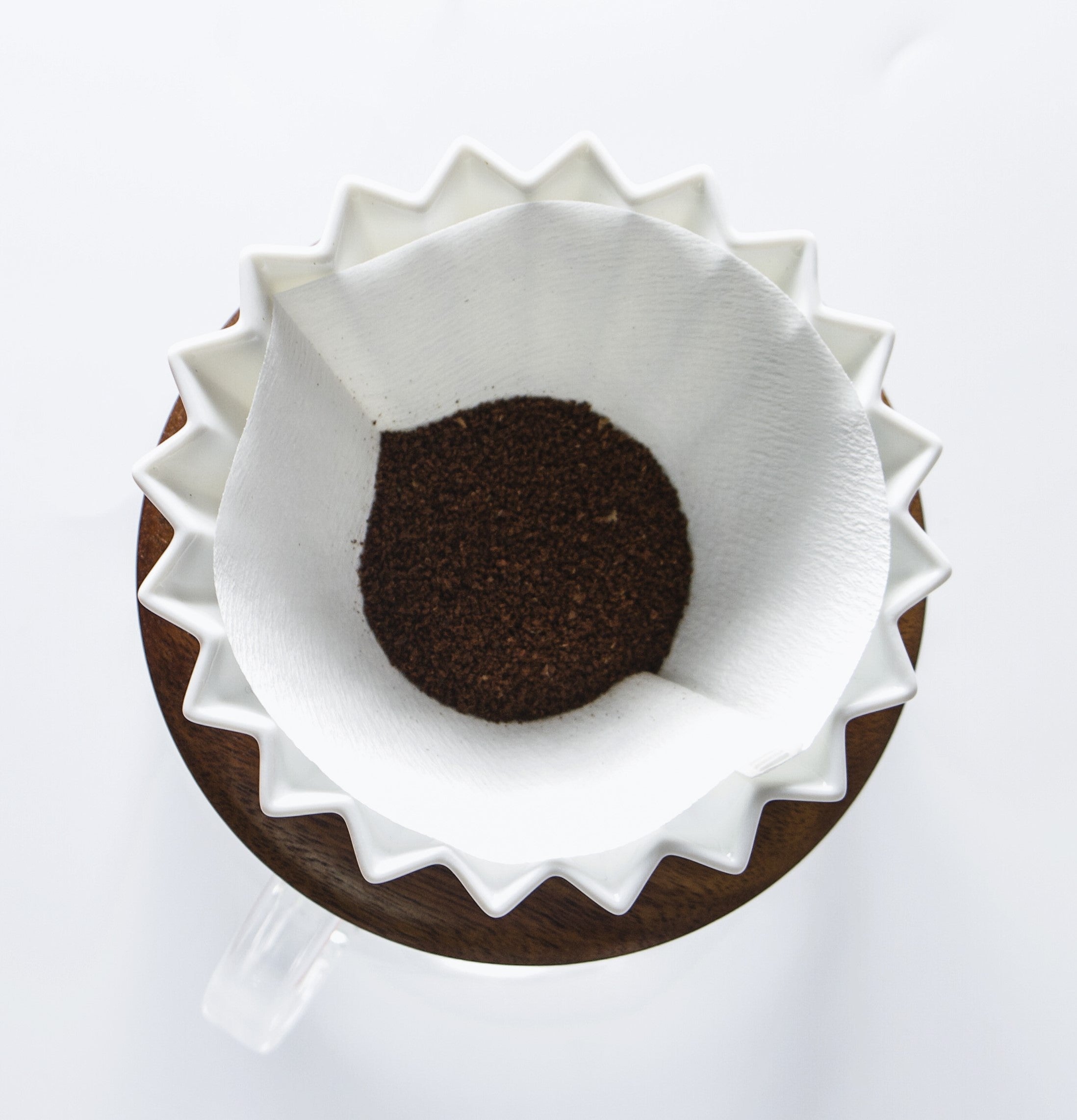 Top-down view of a white Origami coffee dripper with a fluted design, holding a white cone-shaped paper filter filled with ground coffee. The dripper is placed on a wooden base atop a glass server, against a clean white background.