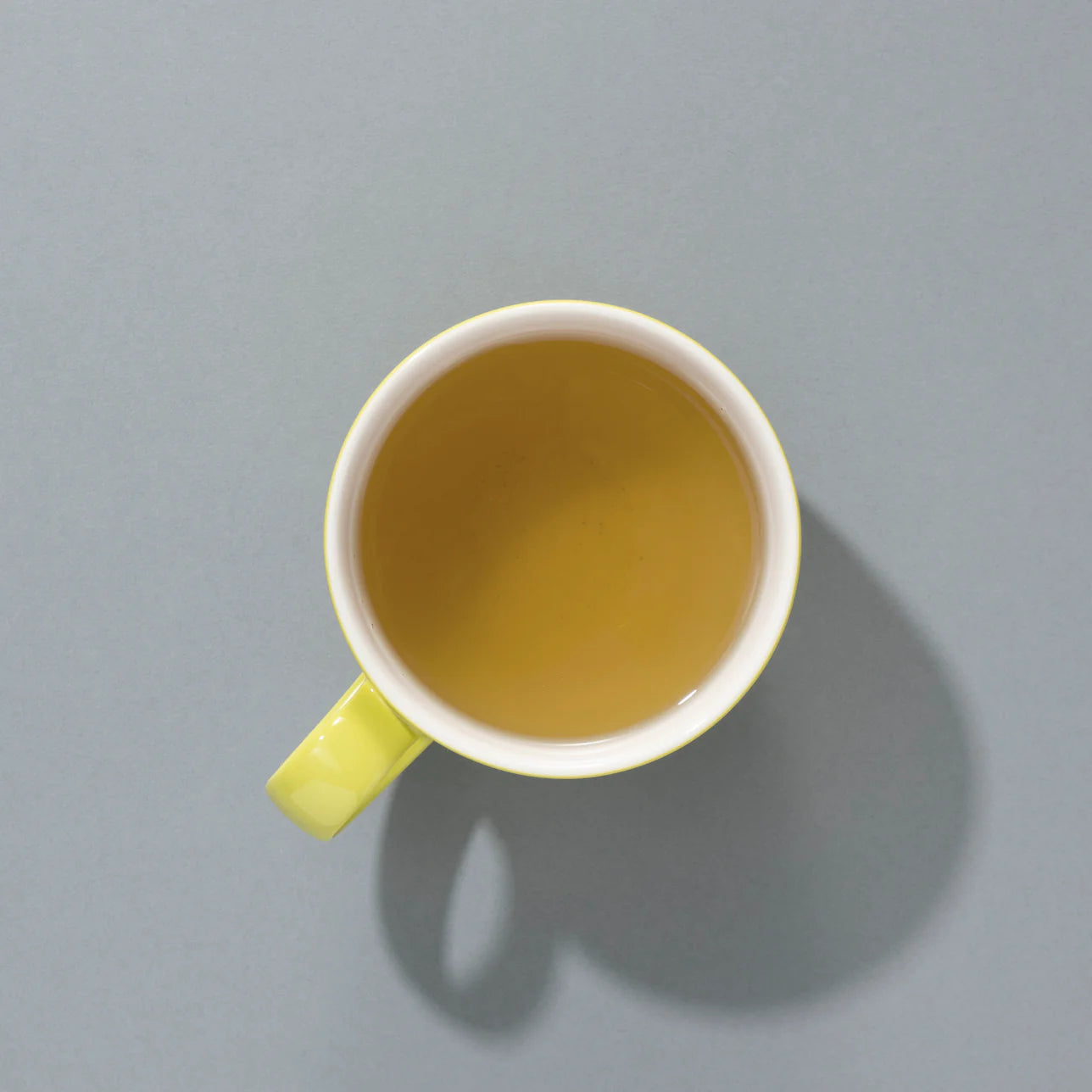 Top-down view of a cup of brewed green Sencha tea in a pale yellow mug, casting a soft shadow on a grey background.