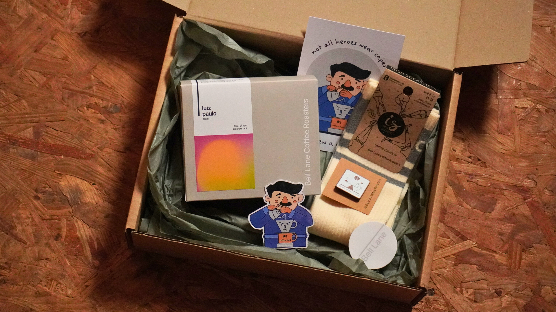 Flat lay of Bell Lane Coffee Roasters’ Father’s Day gift box, featuring a bag of Luiz Paulo coffee, striped socks by Irish Sock Society, a "coffee dad" character postcard and stickers, a branded enamel pin, and a Bell Lane logo sticker, arranged neatly on a vintage leather suitcase.






