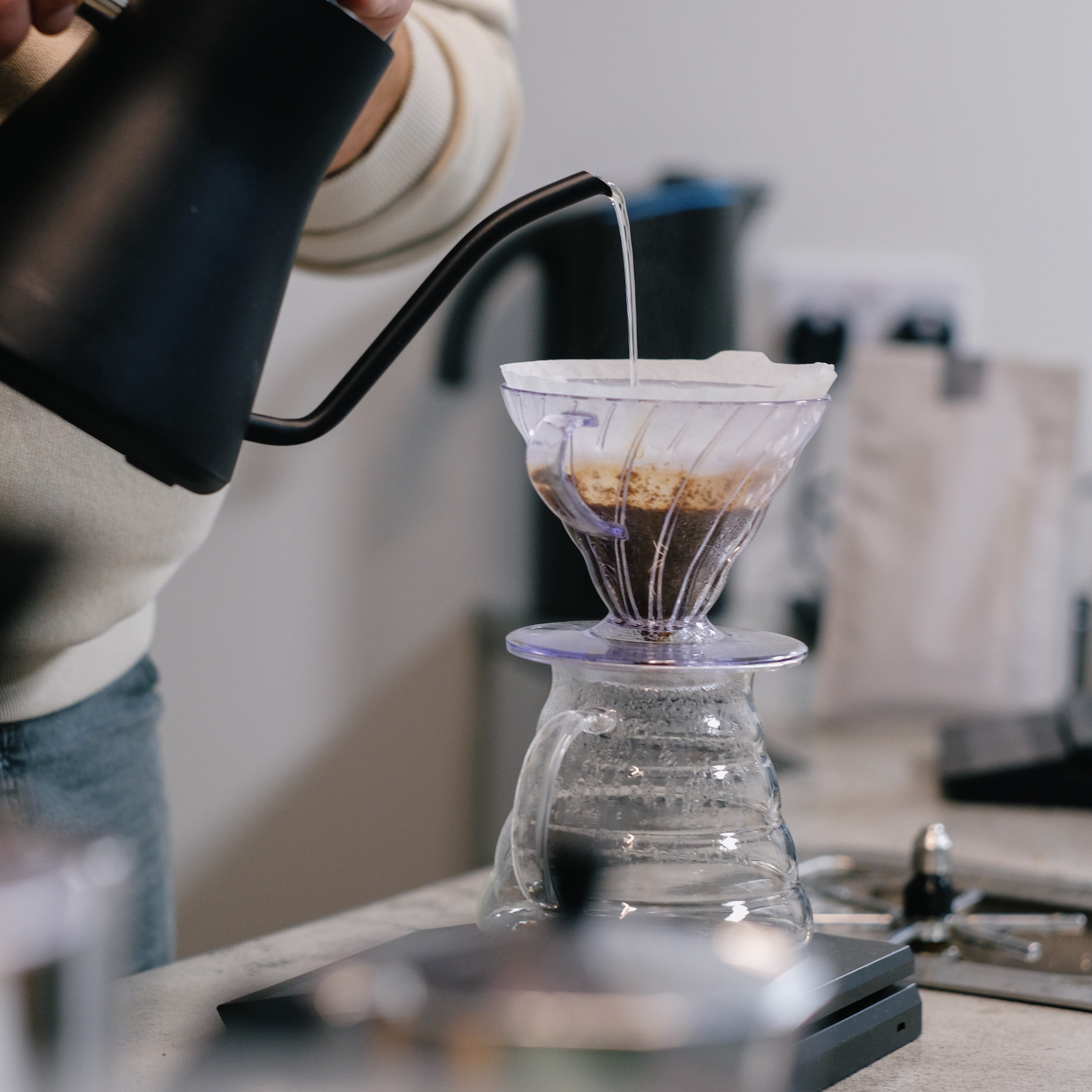 A person is pouring hot water from a black gooseneck kettle into a clear, fluted dripper filled with coffee grounds and a paper filter. The dripper sits atop a glass carafe, which is placed on a digital scale on a countertop. The background includes blurred coffee bags and brewing equipment, suggesting a café or home brewing setup.