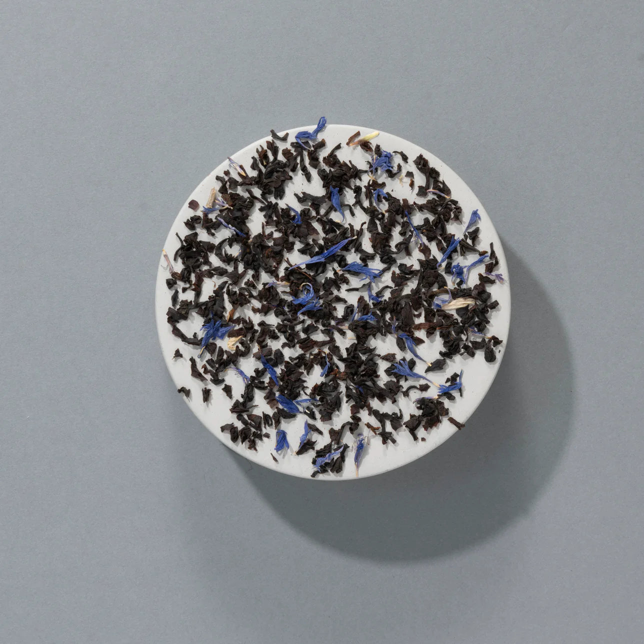 Loose Earl Grey Blue Flower tea leaves with vibrant blue cornflower petals, displayed on a white dish against a grey background.
