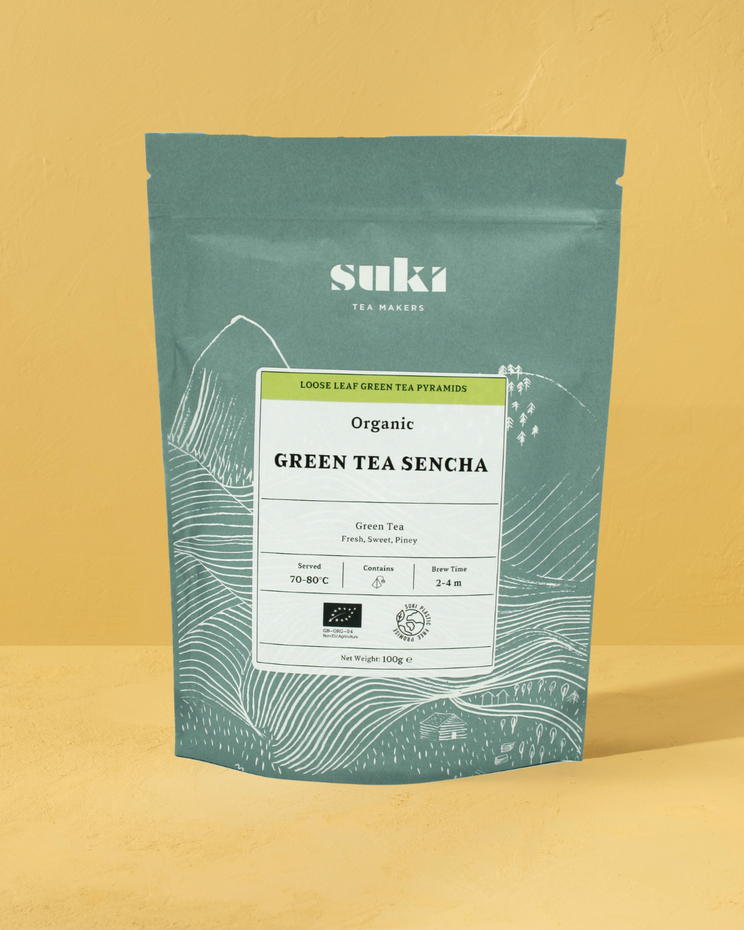 Suki Tea Makers Organic Green Tea Sencha in green plant-based packaging, displayed against a warm yellow background.