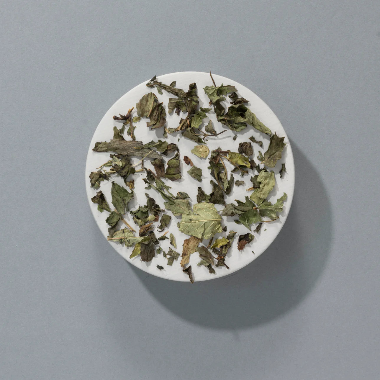 Loose peppermint leaves displayed on a round white dish, set against a soft grey background.
