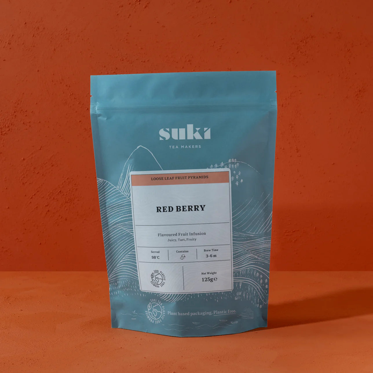 A resealable pouch of Suki Tea Makers Red Berry fruit infusion, displayed against a textured orange background. The blue packaging features a white label with product details and icons indicating temperature, brew time, and certifications.
